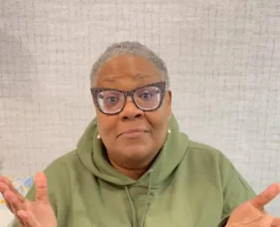 Woman in green hoodie and glasses with short gray hair expressing confusion with raised hands