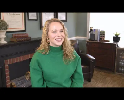Smiling woman with curly blonde hair wearing a green sweater sitting in a cozy office room.