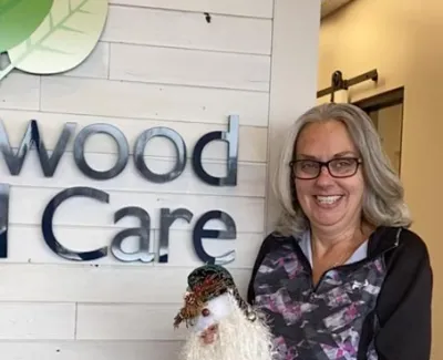 Smiling woman with glasses holding a festive doll standing in front of a wood care company sign.