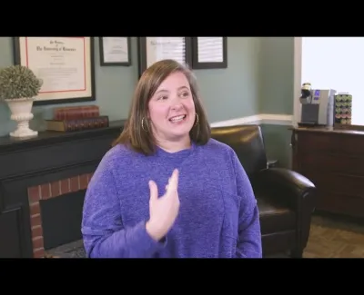 Smiling woman in a blue sweater speaking in a cozy office with framed certificates and leather chair.
