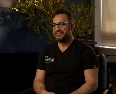 Man wearing glasses and black scrubs sits smiling in a warmly lit modern office with plants.