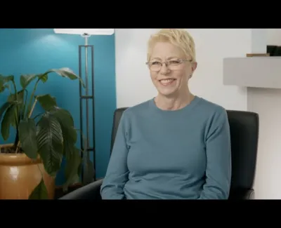 Smiling elderly woman with short blonde hair and glasses sitting on a black chair in a modern room with blue wall and plant.