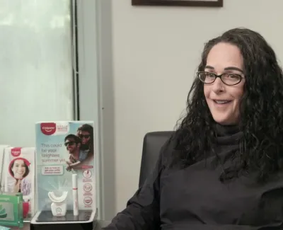 Woman with glasses and curly hair smiles seated in a dental office with product displays and a green informational poster