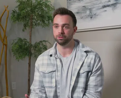 Man in a plaid shirt sitting indoors with decorative plants and abstract art in the background.