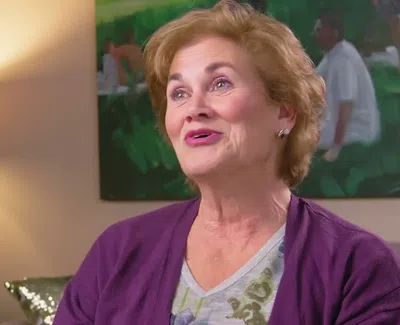 Older woman with short hair wearing purple cardigan speaking in a cozy, art-filled living room with soft lighting.