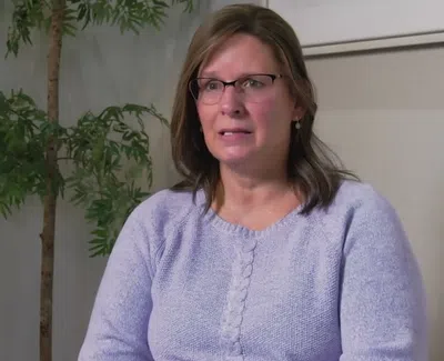 Woman in glasses and light purple sweater sitting indoors with plants in the background