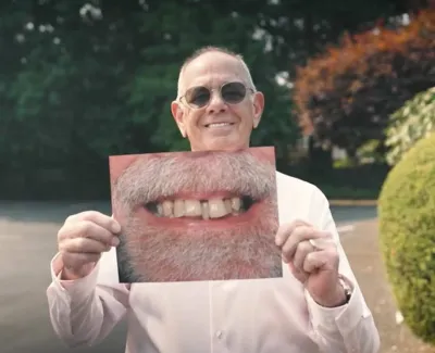 Smiling man wearing sunglasses holds photo of a bearded mouth with missing teeth outdoors in a park.