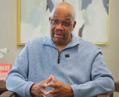 Man in glasses and blue zip-up sweater speaking while sitting in a modern office with abstract art and lamp.