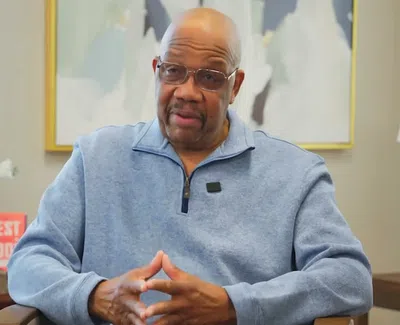 Man in glasses and blue zip-up sweater speaking while sitting in a modern office with abstract art and lamp.