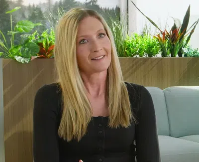 Smiling blonde woman in black shirt sitting on couch with green plants behind her in a bright room