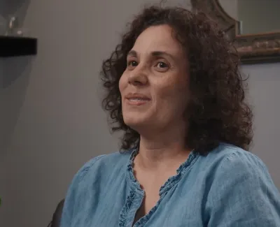 Middle-aged woman with curly hair in a blue blouse sitting indoors near a green plant and speaking.