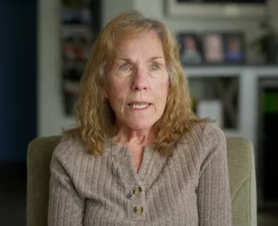 Older woman with curly blond hair wearing a taupe sweater sitting in an indoor living room setting.