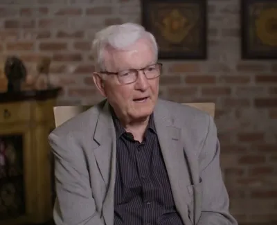 Elderly man with white hair and glasses speaking while seated in a room with brick wall and framed art.