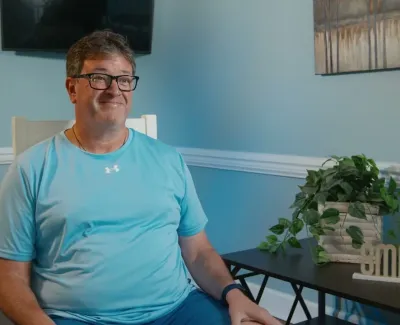 Middle-aged man in blue shirt sitting in a bright room with plants and a smile decoration on a table beside him