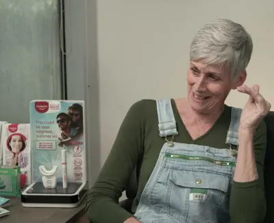 Mature woman with short gray hair smiling while sitting in a dental office with promotional displays.