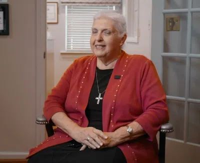 Elderly woman with short white hair wearing red cardigan and cross necklace, seated indoors in an office setting.