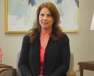 Woman with brown hair in a black cardigan and coral shirt sits in a bright office setting with awards and a lamp.