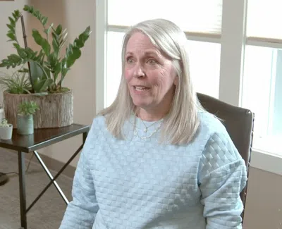 Smiling woman with gray hair sitting indoors near window and table with potted plants in a cozy living room.