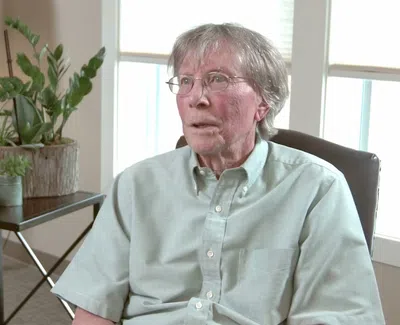 Elderly man with glasses sitting in a light-filled room next to a table with potted plants and a lamp.