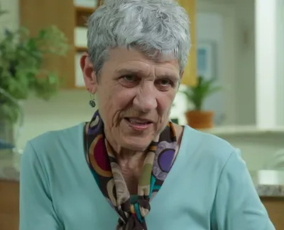 Elderly woman with gray hair wearing a colorful scarf and turquoise top in a bright, cozy room.