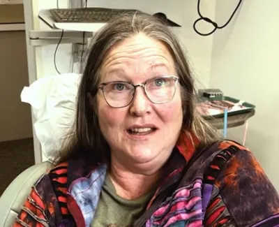 Middle-aged woman with glasses wearing a colorful jacket sitting indoors with medical equipment in background