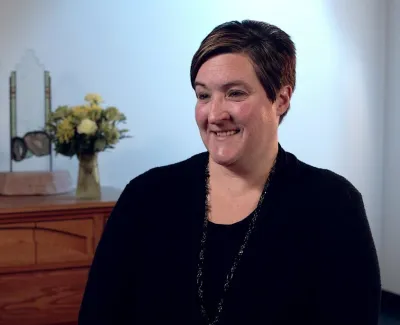 Smiling woman with short hair sitting indoors with wooden dresser, flowers, and decorative items in background.
