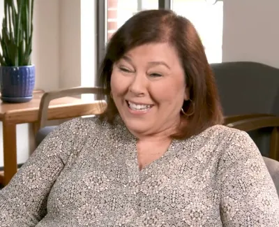 Smiling middle-aged woman seated in a room with wooden furniture and a potted plant in the background