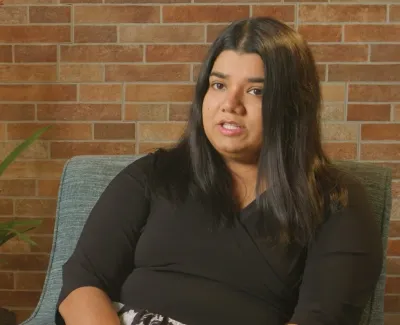 Woman with long dark hair speaking while seated in a chair against a brick wall and potted plants.