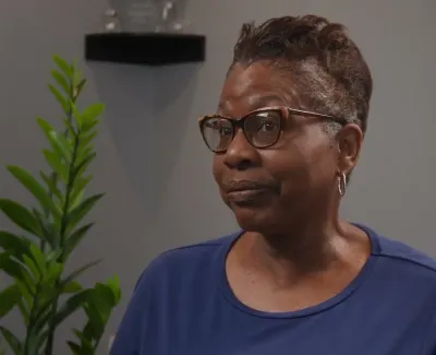 Middle-aged woman with glasses and short hair wearing a blue shirt sitting indoors near green plant and gray wall.