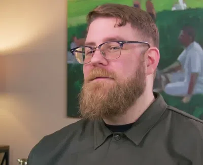 Man with glasses and beard wearing a dark shirt sitting indoors with lamp and painting in the background
