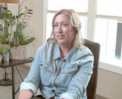 Woman in a light denim shirt sitting and talking in a cozy, plant-filled living room with natural light.