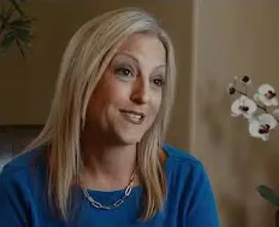 Smiling woman with blonde hair in blue top seated indoors near plants and white orchids.