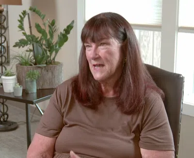 Middle-aged woman with brown hair talking while seated in a bright, cozy living room with plants and furniture.