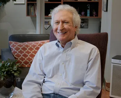 Smiling elderly man sitting in a cozy living room with plants, pillows, and books in the background