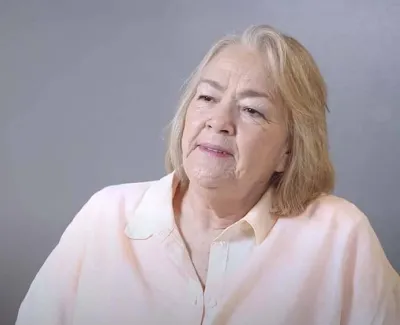 Older woman with blonde hair wearing a light pink shirt against a gray background looking thoughtfully.
