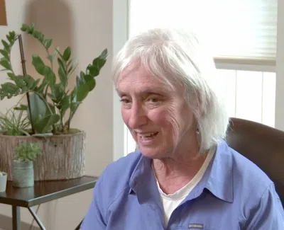 Elderly woman with white hair smiling, sitting in chair near window and indoor plants on table behind her.