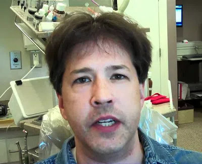 Man with brown hair speaking in a medical or dental office with equipment in the background