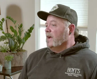Man wearing black Hoyt's hoodie and cap sitting indoors with plants and lamps in the background