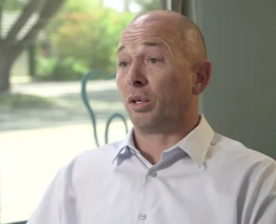 Bald man in a white collared shirt speaking indoors near a window with a blurred outdoor background.