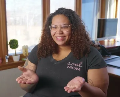 Smiling woman with curly hair and glasses wearing a black Cool Mom t-shirt in a cozy office setting.