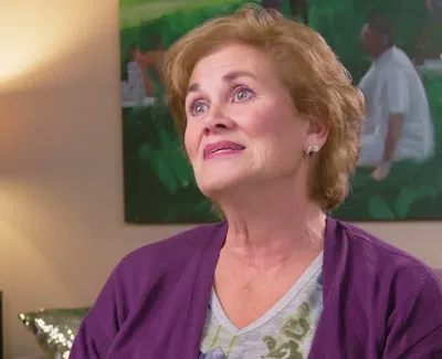 Middle-aged woman with short blonde hair wearing a purple cardigan, sitting indoors with a thoughtful expression.