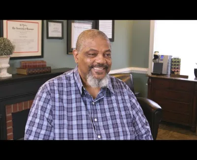 Smiling man with gray beard wearing a blue plaid shirt sitting in a cozy room with certificates and books behind.