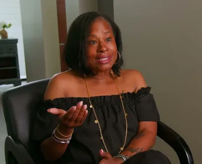 Woman in black off-shoulder top speaking animatedly while seated in a black chair indoors.