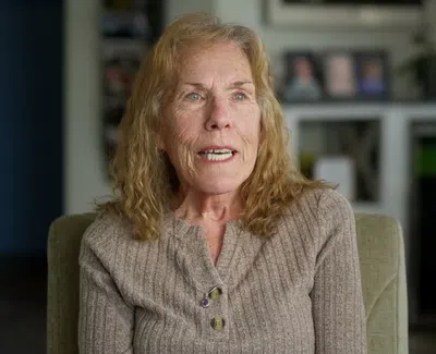 Older woman with curly blonde hair wearing a brown sweater sitting in a cozy room speaking.