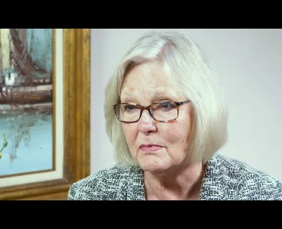Older woman with light hair and glasses in professional attire sitting indoors near a framed painting and plant.