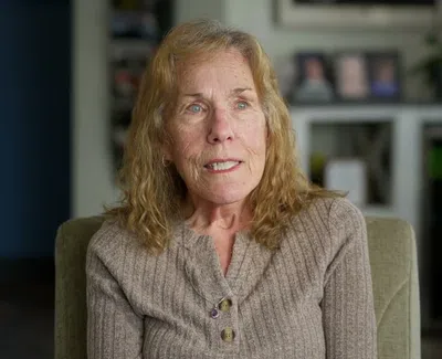 Mature woman with blonde hair wearing a brown sweater sitting in a cushioned chair indoors.