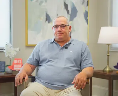 Middle-aged man wearing glasses sitting in a chair in a bright room with award plaques and a lamp.