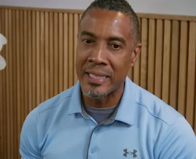 Man in blue Under Armour polo speaking indoors with white orchids and wooden panel background