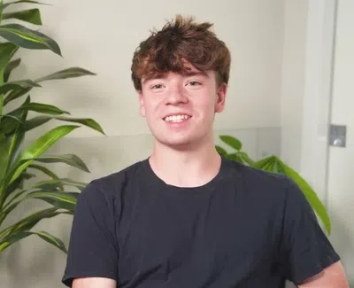 Smiling young man with brown hair wearing a black t-shirt sitting indoors with green plants in the background.