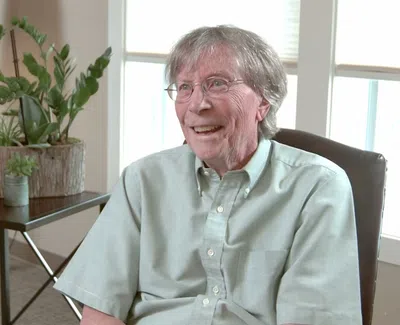 Elderly man smiling and seated in a bright room with indoor plants and a window in the background.
